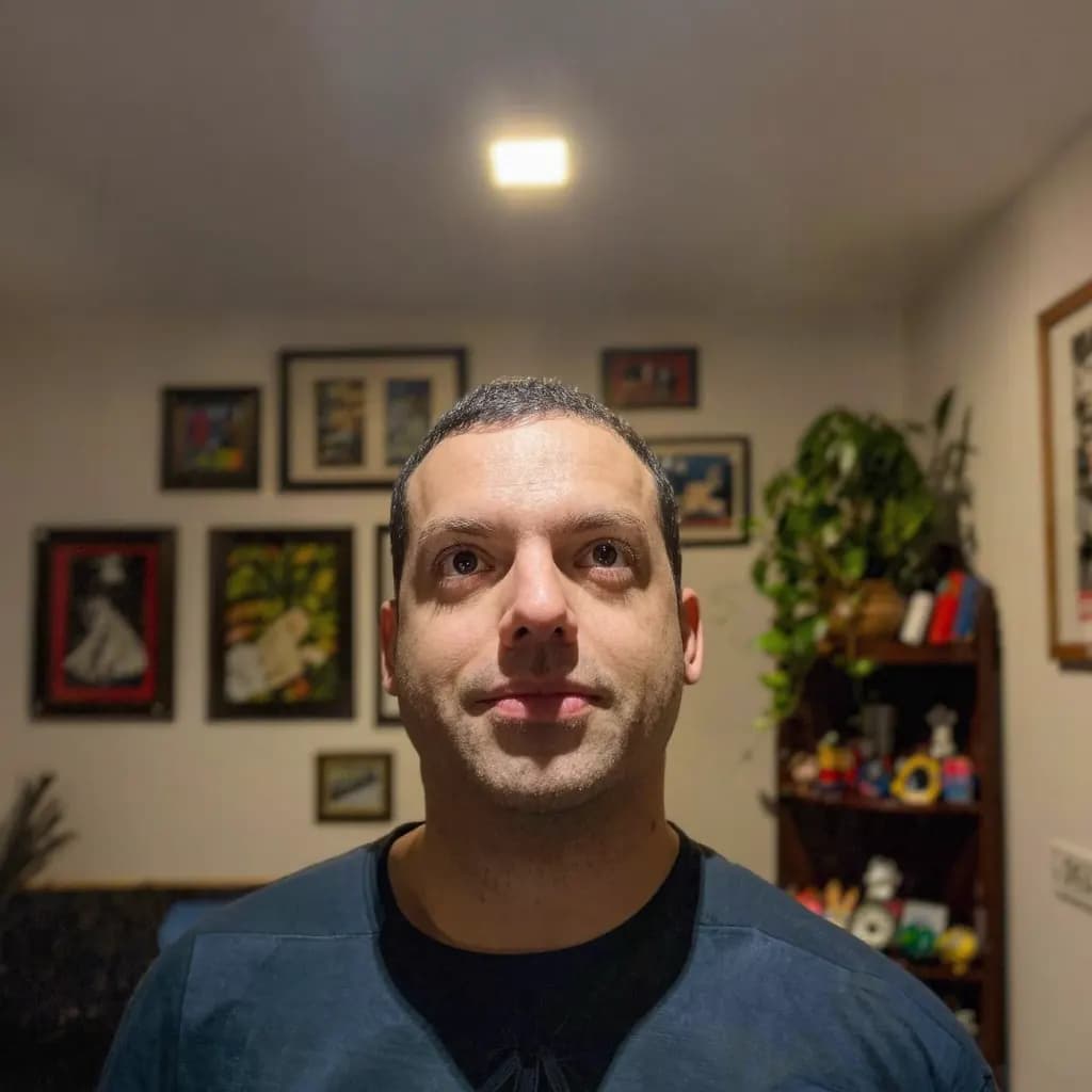 A man lit by a ceiling lamp, with shadows under his eyes and pictures on the wall behind