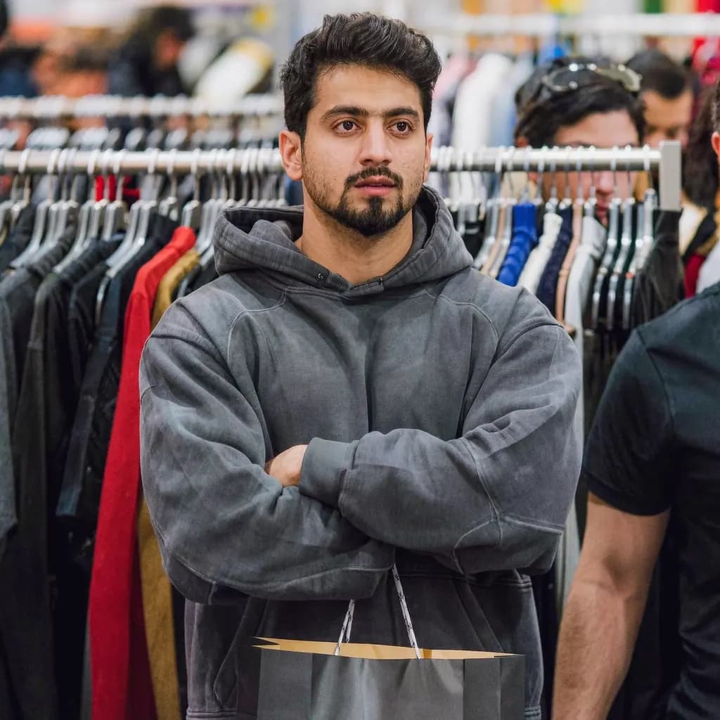 A man cropped at the knees with his arms crossed, in a baggy hoodie in a busy shop